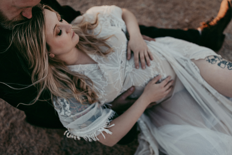 Maternity Photographer, a pregnant woman lays in her partner's arms outside as they recline on rock. She wears a white dress.