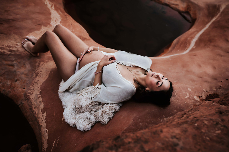 Maternity Photographer, a pregnant woman in a white dress lays on red desert rock